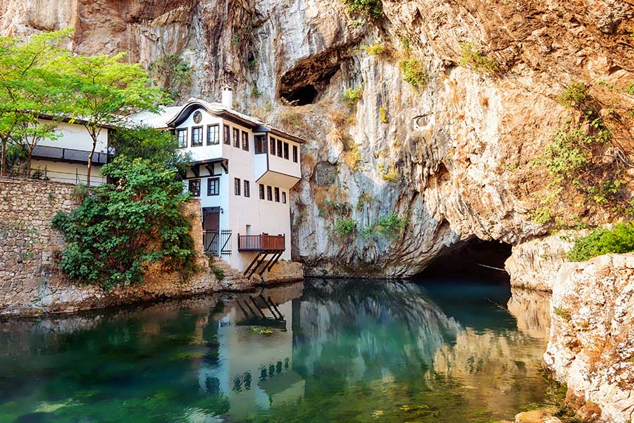 Ein weißes Haus steht unter einer Felswand, davor ein Fluss, der aus einer Höhle kommt