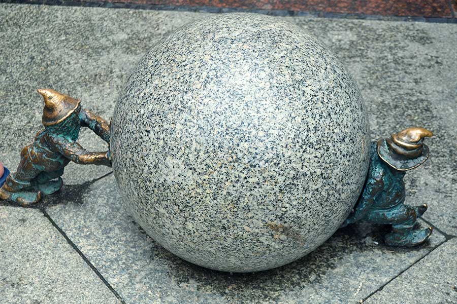 Zwei Bronzezwerge schieben eine Steinkugel von entgegengesetzten Seiten