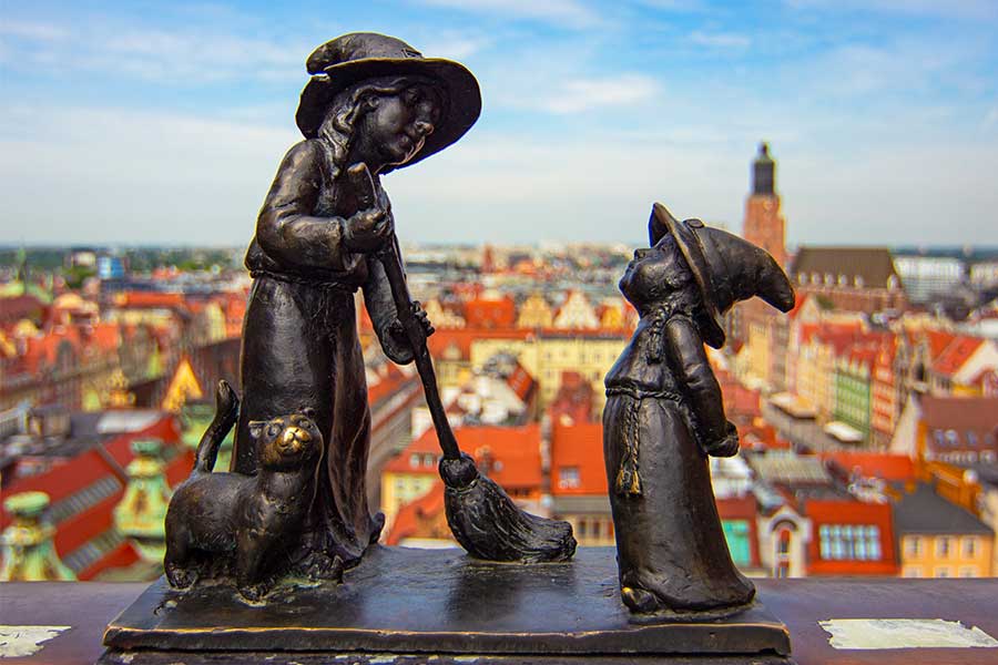 Bronzeskulptur aus kleiner und großer Hexe mit Besen und Katze, dahinter Blick über die Dächer von Breslau